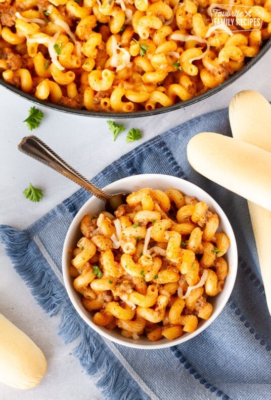 Small bowl of Creamy Ground Beef and Noodles Skillet with a fork next to a skillet of Creamy Ground Beef and Noodles. Bread sticks on the side.