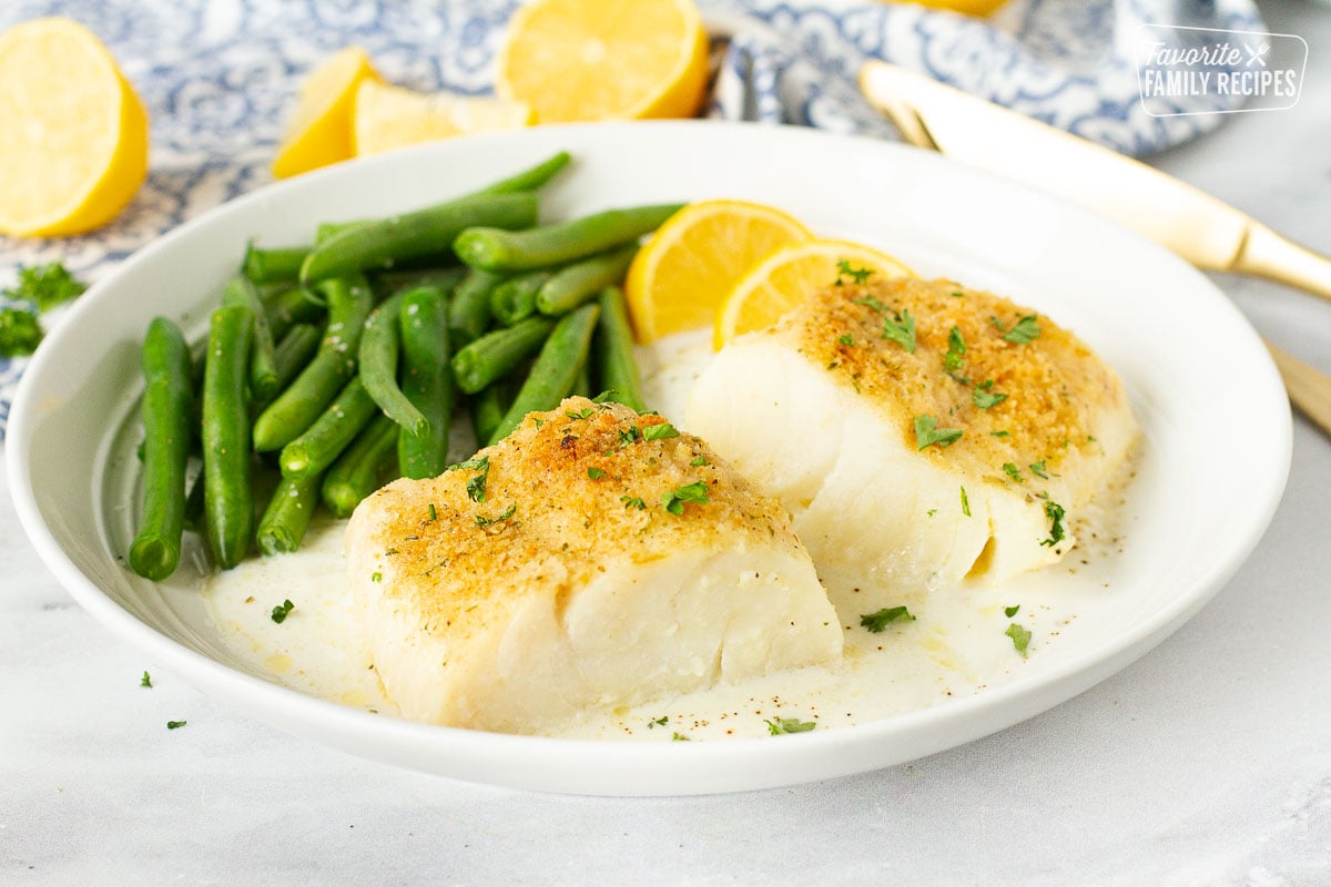 Plate with two fillets of Baked Cod in Cream Sauce with green beans and lemon wedges.