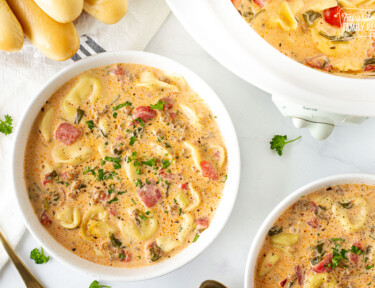 Crockpot of Creamy Tortellini Soup next to two bowls of Creamy Tortellini Soup and breadsticks.