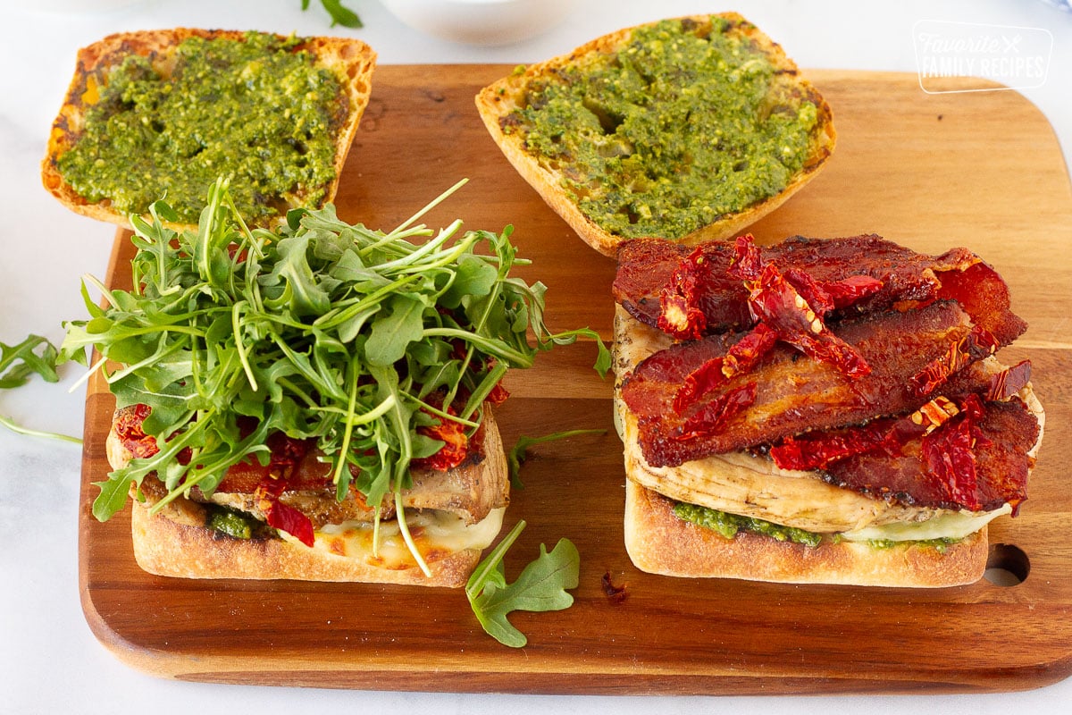 Assembling two Chicken Pesto Sandwiches on a cutting board with ciabatta bread, pesto mayo, chicken breast, bacon, sun dried tomatoes and arugula.