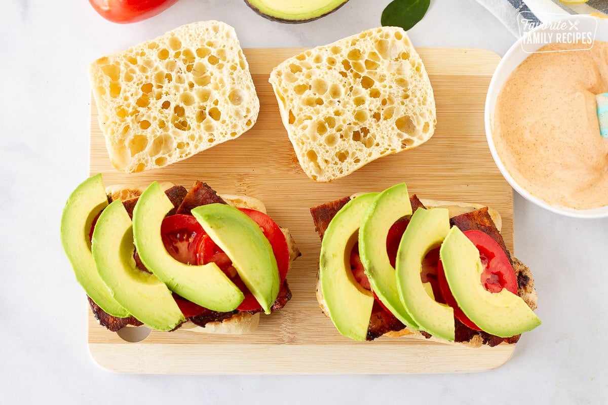 Cutting board with ciabatta bread topped with chicken, bacon, tomatoes and avocado for Grilled Chicken Sandwiches.