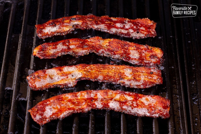 Four strips of bacon grilling on the barbecue for Grilled Chicken Sandwiches.