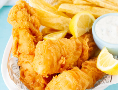 Basket of three Fish and Chips with a dipping bowl of tartar sauce and lemon wedges.