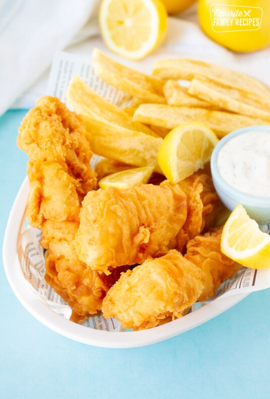 Basket of three Fish and Chips with a dipping bowl of tartar sauce and lemon wedges.