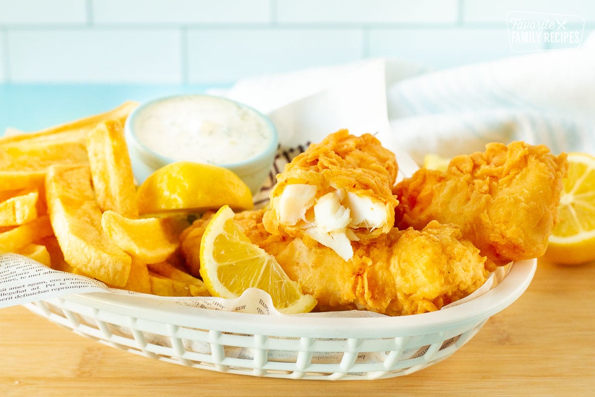 Inside view of one fish fillet in a basket of Fish and Chips. Lemon wedges and tartar sauce on the side.