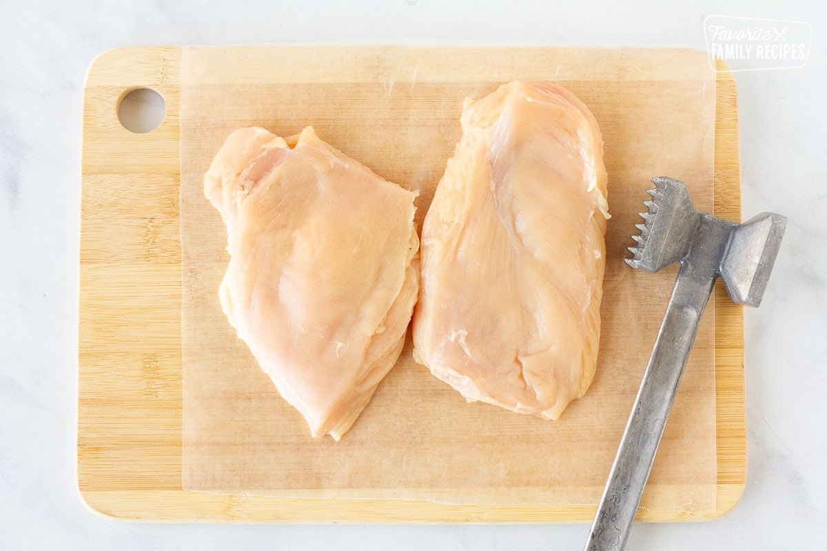 Cutting board with flattened chicken breast halves and metal mallet for Grilled Chicken Sandwiches.