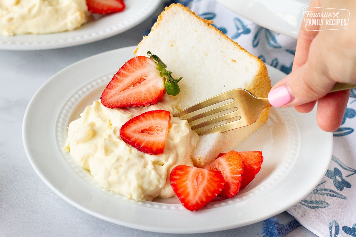 Hand holding a fork cutting into a slice of Angel Food Cake with pineapple whipped topping and fresh sliced strawberries.