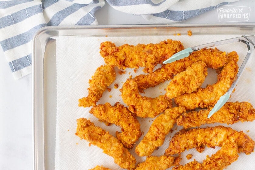 Fried chicken on a cookie sheet with a paper towel and tongs for Applebee's Oriental Chicken Salad.