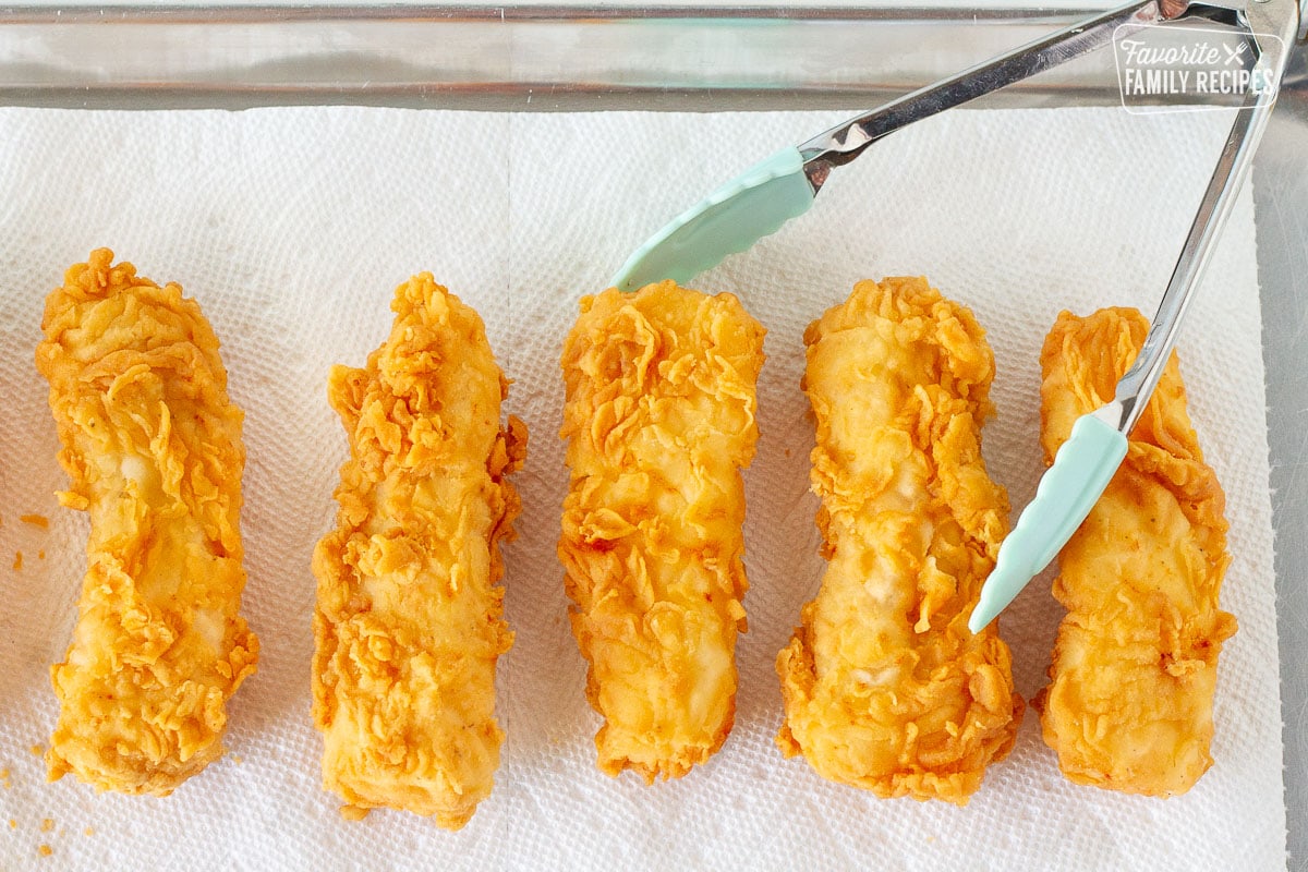 Five fried Cod fillets on paper towels for Fish and Chips. Tongs on the side.