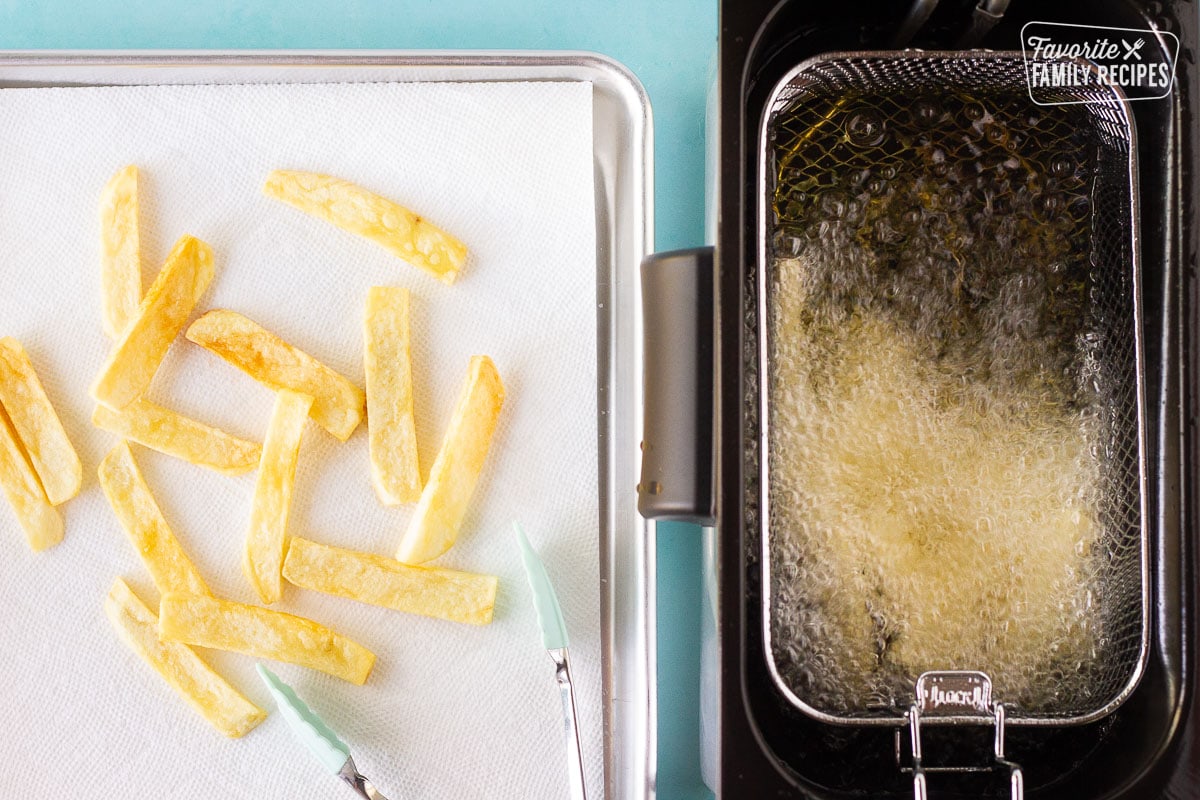 Frying cut up potatoes in the fryer next to a baking sheet with French fries and tongs for Fish and Chips.