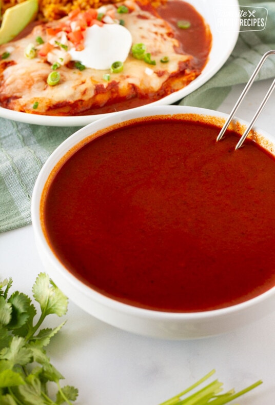 Bowl of Homemade Enchilada Sauce with a ladle resting inside next to a plate of cheese enchiladas.