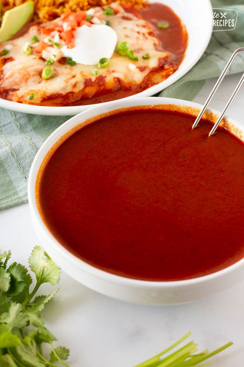 Bowl of Homemade Enchilada Sauce with a ladle resting inside next to a plate of cheese enchiladas.