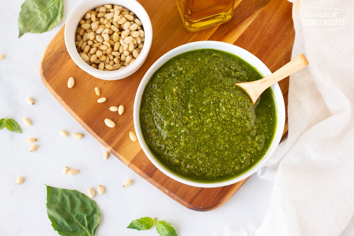 Homemade Pesto in a bowl with a wooden spoon next to a bowl of pine nuts and olive oil.