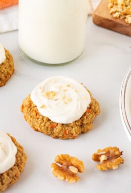 Carrot Cake cookies with cream cheese frosting on a plate and on the table. Milk and walnuts on the side.