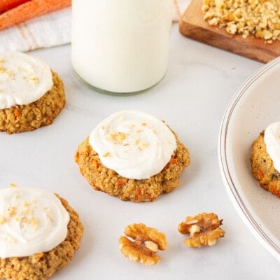 Carrot Cake cookies with cream cheese frosting on a plate and on the table. Milk and walnuts on the side.