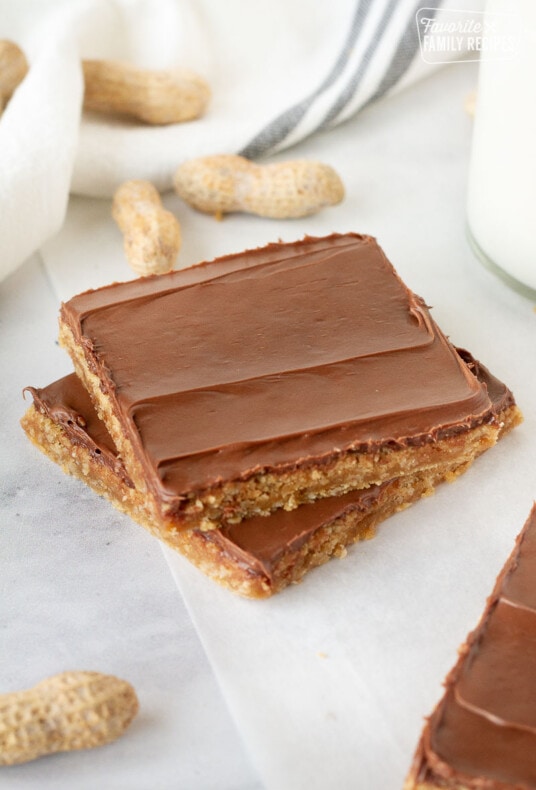 Two Peanut Butter Bars stacked together next to milk. Extra whole peanuts on the side.