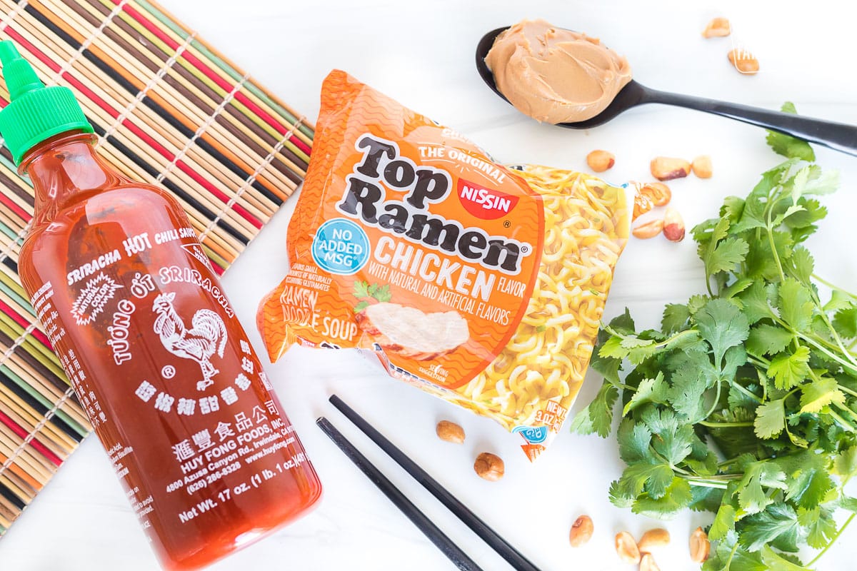 A package of ramen next to a spoon of peanut butter and a bottle of sriracha