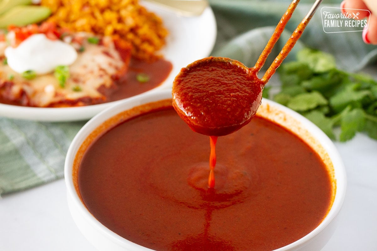 Pouring enchilada sauce with a ladle into a bowl. Plate of homemade enchiladas in the background.