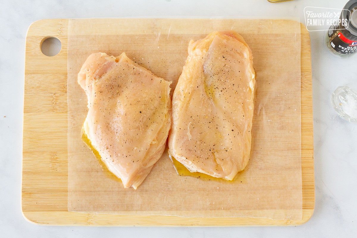 Cutting board with flattened chicken breasts with oil, salt and pepper for Chicken Pesto Sandwiches.