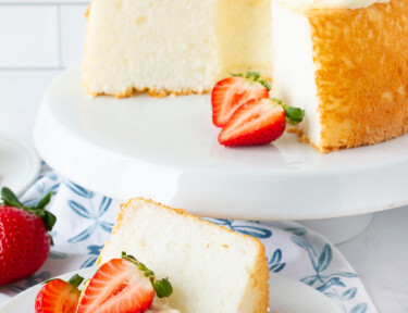 Angel Food Cake with pineapple whip topping and fresh sliced strawberries on a cake stand next to a plate with a slice of Angel Food Cake.