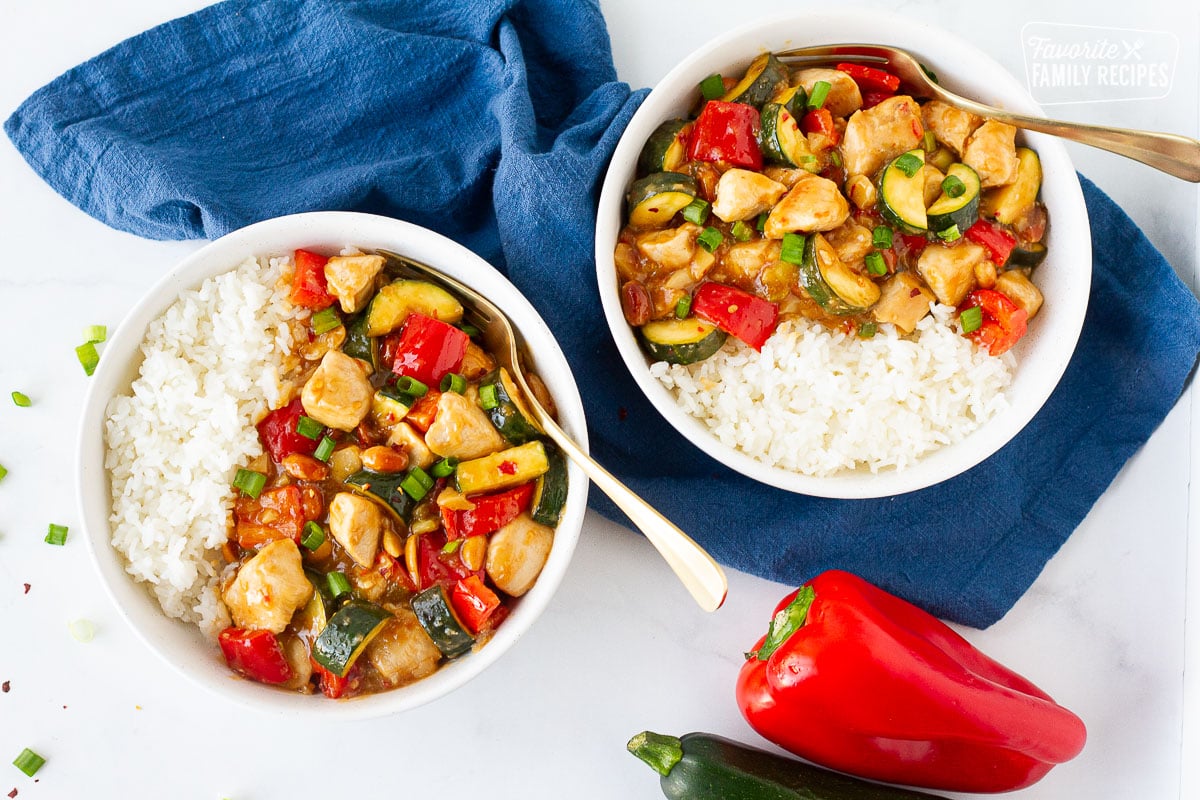 Two bowls of Kung Pao Chicken with white rice. Red bell pepper and zucchini on the side.