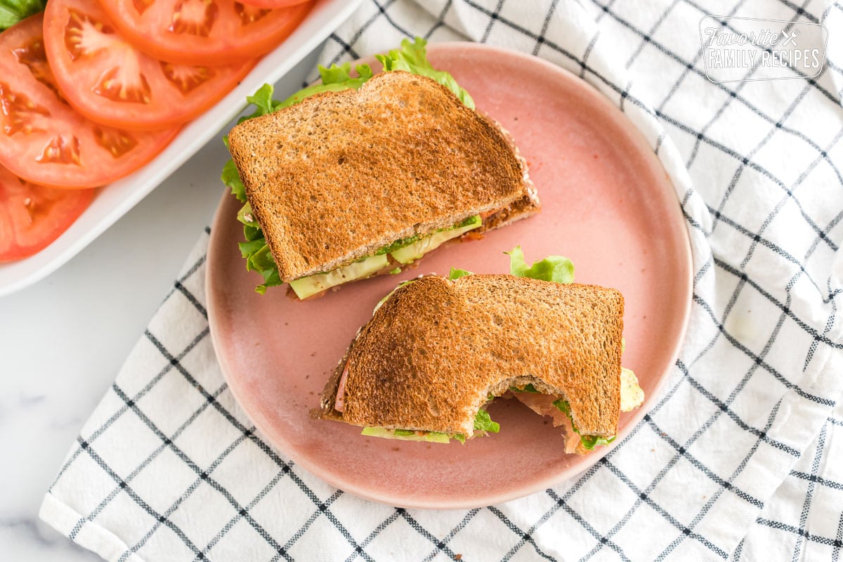 Ultimate BLT cut in half and a bite taken out of it on a pink plate. 