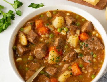 Hearty Beef Stew in a bowl with a spoon. Buttered dinner roll on the side.