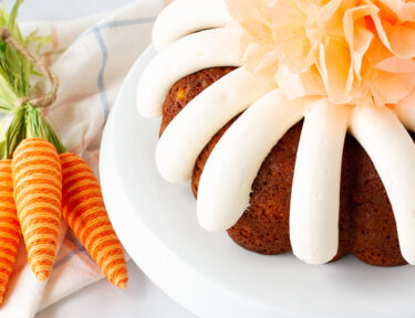 Angle view of Carrot Bundt Cake on a cake stand with a decorative tissue pom pom in the center. Plates and forks on the side.