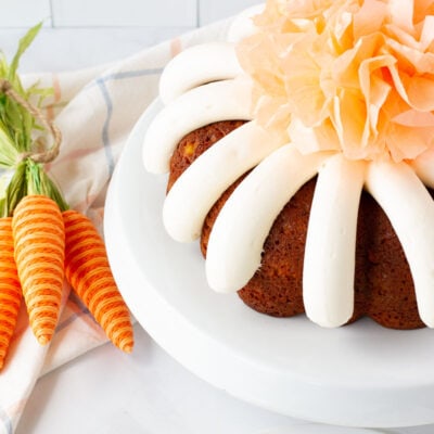Angle view of Carrot Bundt Cake on a cake stand with a decorative tissue pom pom in the center. Plates and forks on the side.