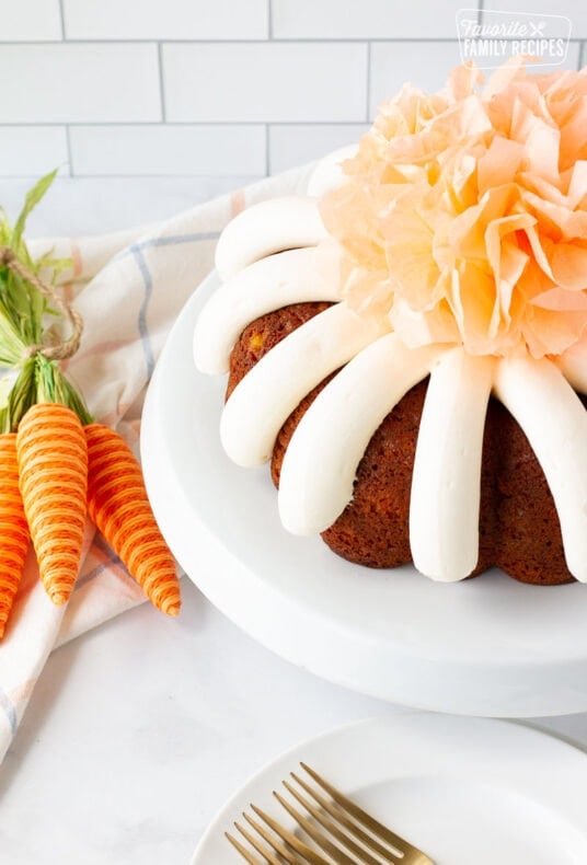 Angle view of Carrot Bundt Cake on a cake stand with a decorative tissue pom pom in the center. Plates and forks on the side.