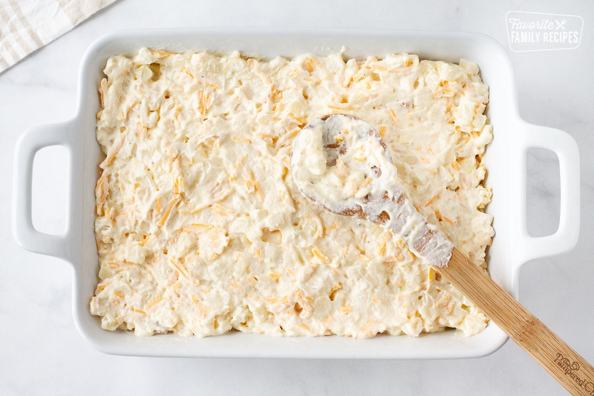Cheesy Potato Casserole mixture in a baking dish spreading with a wooden spoon.