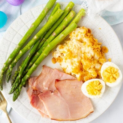 Sliced ham, asparagus spears, cheesy potato casserole, and deviled eggs on a Dinner Plate for Easter Ideas. Rolls and Easter eggs on the side.