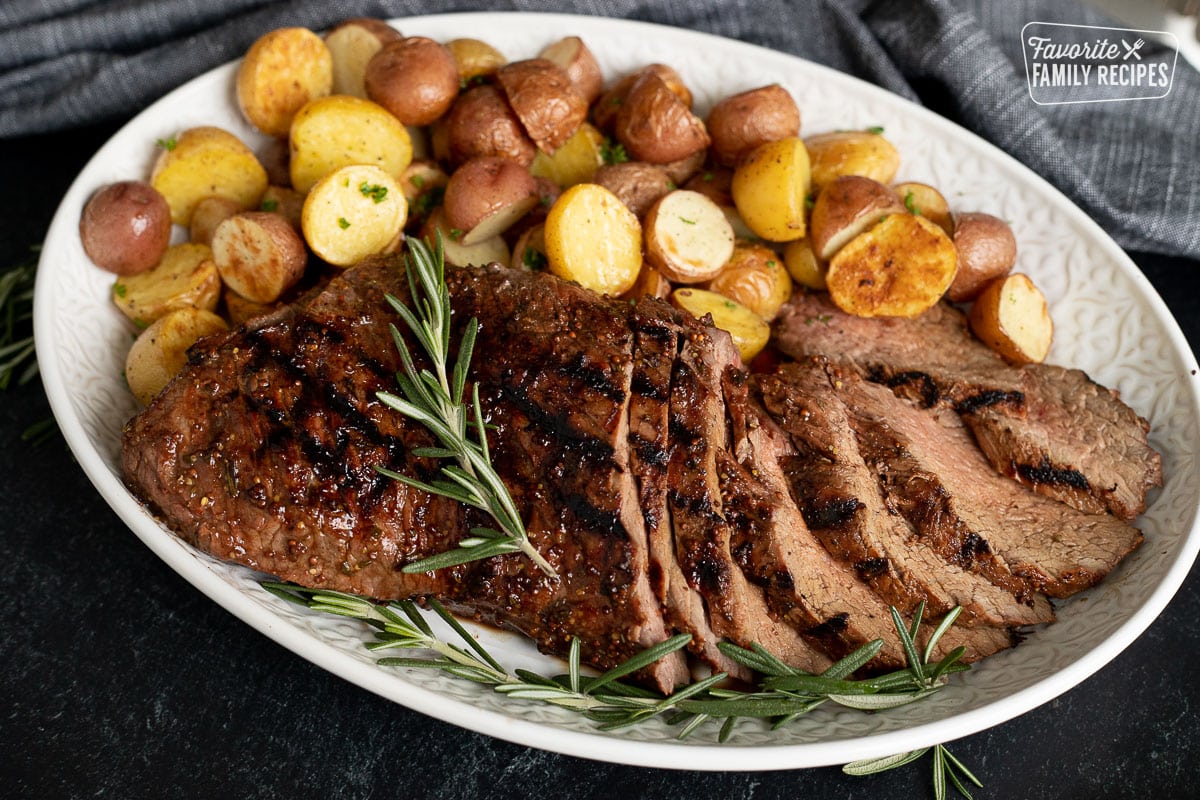 Sliced Tri Tip Roast garnished with fresh rosemary on a tray with red and golden potatoes.