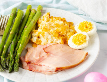Side view of an Easter Dinner Ideas with cheesy potato casserole, asparagus, eggs and ham. Bowl of rolls on the side.