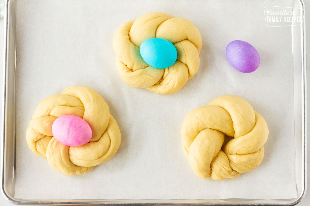 Baking sheet lined with parchment paper with three Easter Bread round loaves and a colored egg on top in the center of the twisted loaf.