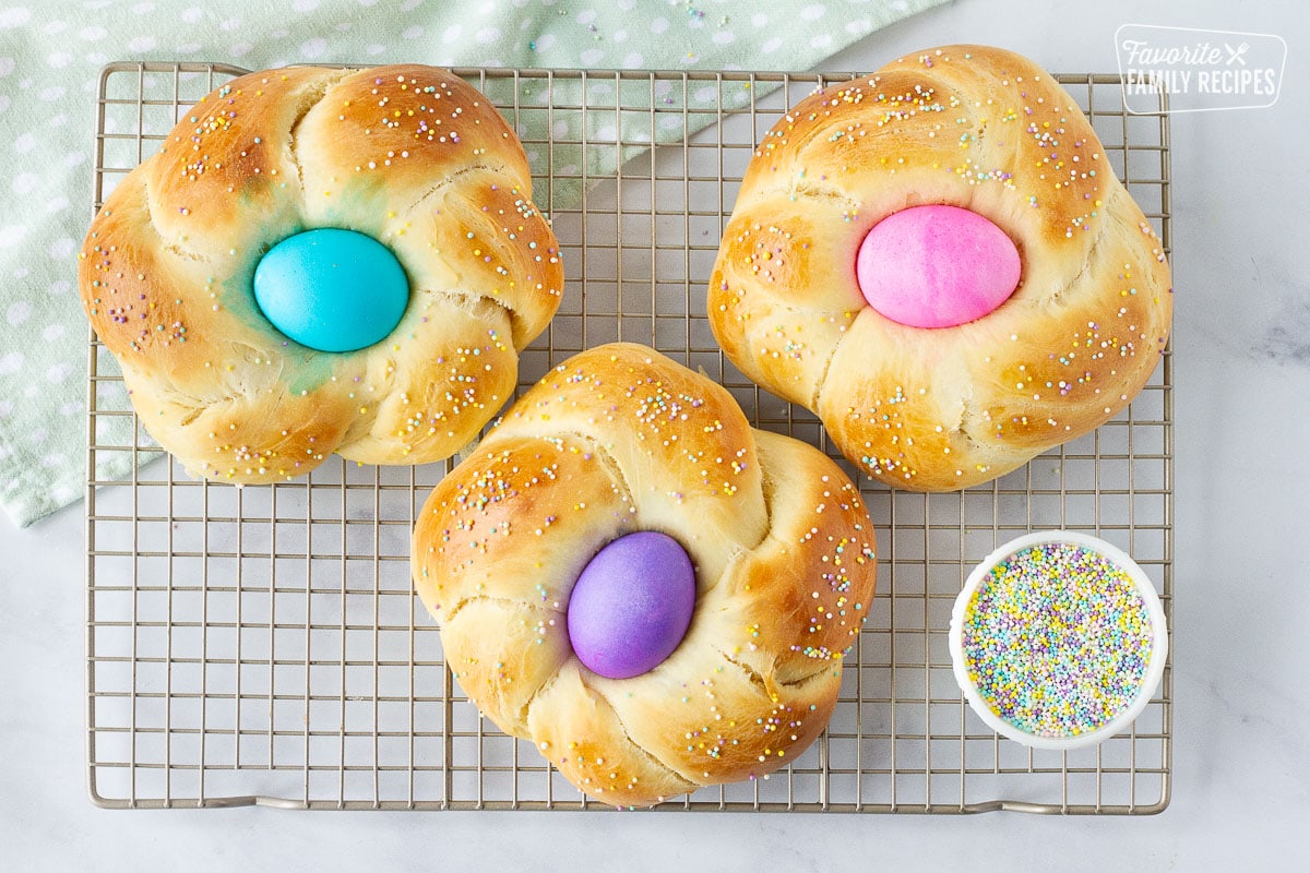 Three loaves of baked Easter Bread cooling on a wire rack and a mini bowl of sprinkles.