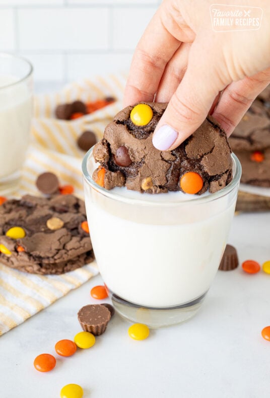 Hand dipping a Reeses Pieces Cookie in a glass of milk.