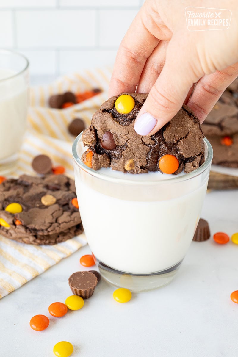 Hand dipping a Reeses Pieces Cookie in a glass of milk.