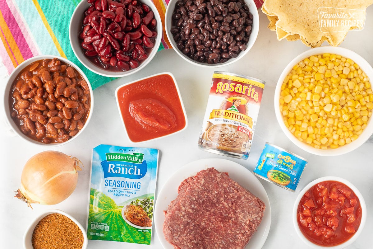 Ingredients to make Mexican Chili including Mexican Chili Beans, kidney beans, black beans, tomato sauce, corn, refried beans, onion, ranch seasoning, taco seasoning, diced tomatoes, ground beef, diced chilis and tostada bowls.