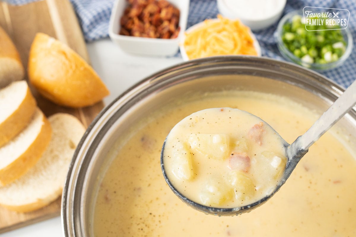 Ladle with Loaded Potato Soup over a pot. Sliced bread, bacon, cheese, onions and sour cream on the side.