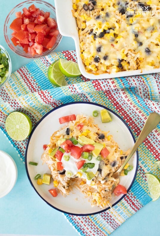 Baking dish of Mexican Chicken Casserole next to a plate with a slice of Mexican Chicken Casserole topped with sour cream, avocado, green onions and tomatoes.