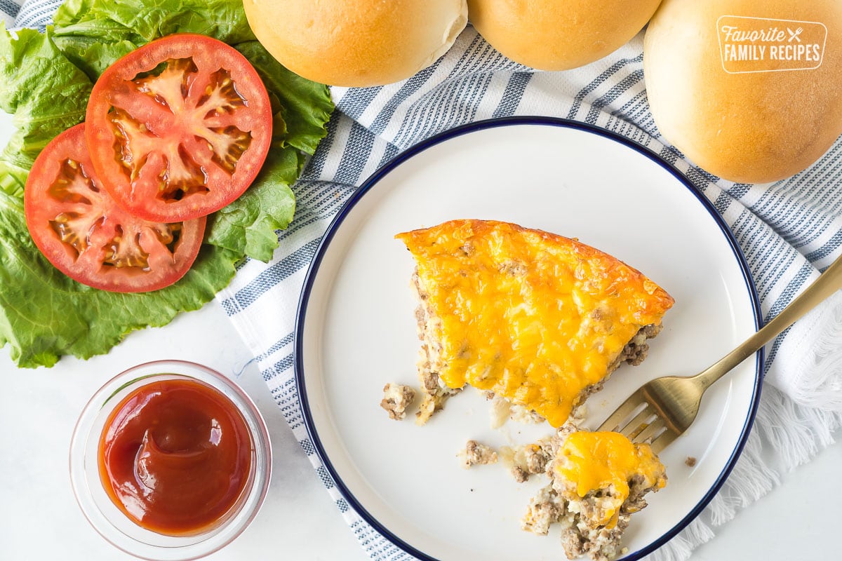 Cheeseburger Pie slice with a fork. Ketchup, lettuce and tomatoes on the side served with buns.