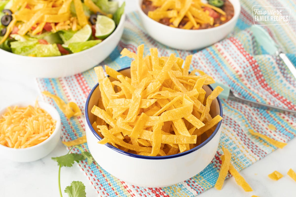 Bowl of Tortilla Strips next to a salad and bowl of chili garnished with Tortilla Strips.