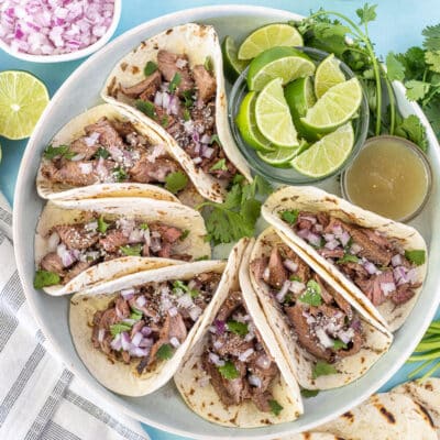Street tacos on a plate with limes, cilantro and red onions.