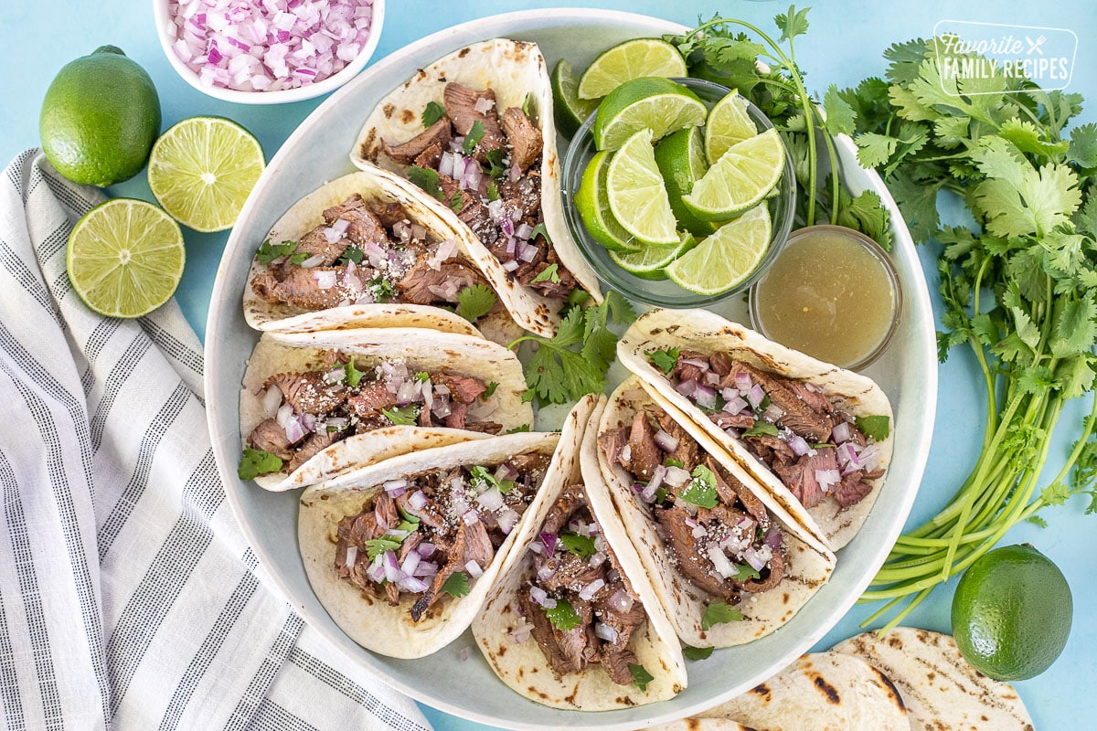 Street tacos on a plate with limes, cilantro and red onions.