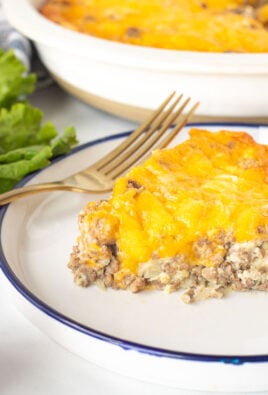 Slice of Cheeseburger Pie on a plate with lettuce, tomatoes and rolls on the side.