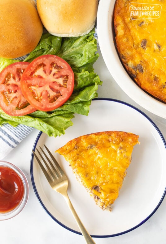 Slice of Cheeseburger Pie on a plate with ketchup, sliced tomatoes, lettuce and buns on the side.