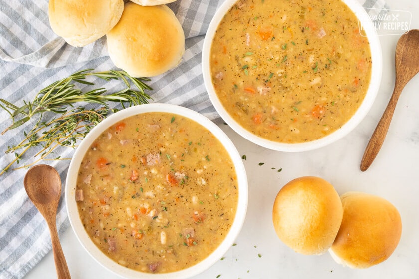Two bowls of Ham and Bean soup with fresh herbs, spoons and rolls on the side.