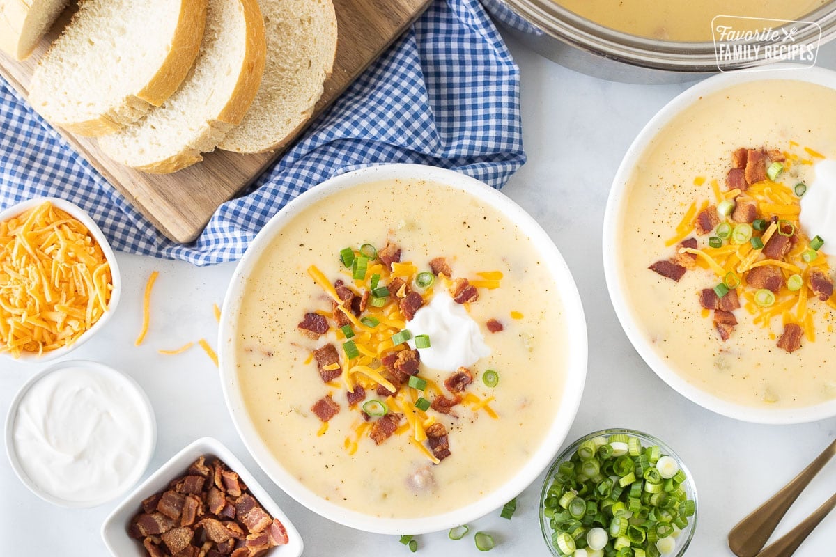 Whether you're looking for a new go-to soup, or just want something creamy and comforting, this Cheesy Potato Soup recipe is sure to satisfy! It is loaded with so much flavor. Two bowls of Loaded Potato Soup with bread. Bacon, cheese, sour cream, onions on the side.