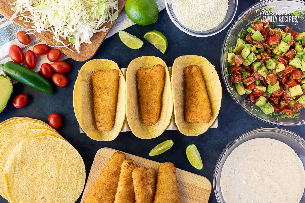 Three corn tortilla shells each with a cooked breaded fish fillet. Pico de Gallo, cilantro cream sauce, cabbage and limes on the side.